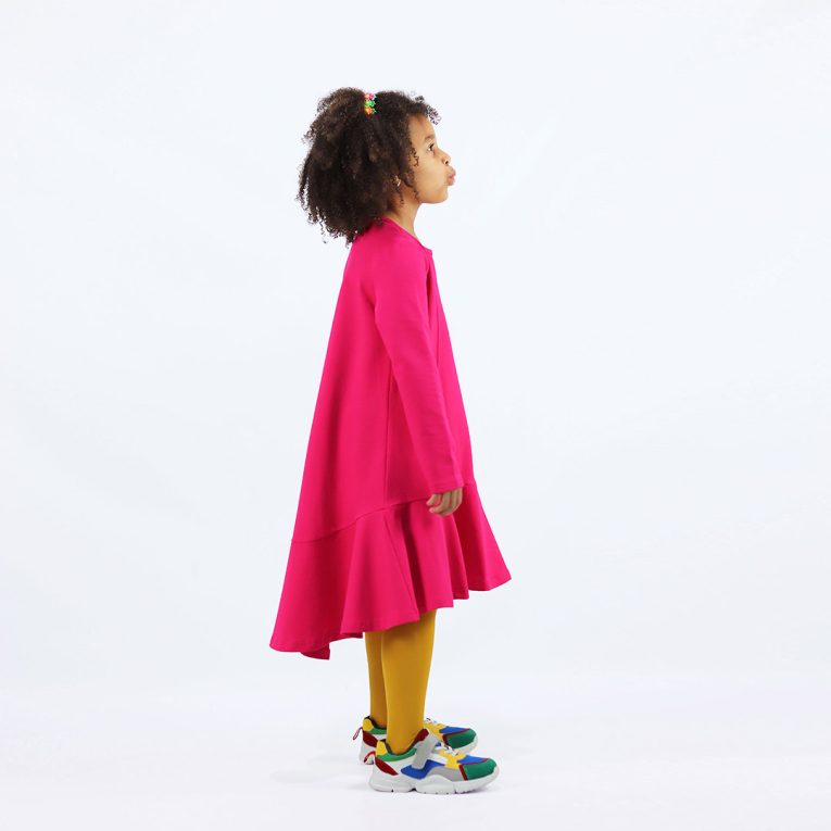 Loose dress is a loose fit dress, that goes wider to the bottom. It is longer on the back with a wide frilled trim at the bottom. Dress is in raspberry colour. Children, 3 -10 yrs. BonnyJoy - side view