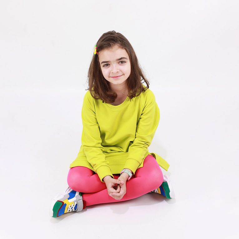 Cylinder dress is a one-colour loose fit dress with round pockets aside and a little longer on the back in bright lime colour. Front view, a girl sitting. Children, 3 -10 yrs. BonnyJoy
