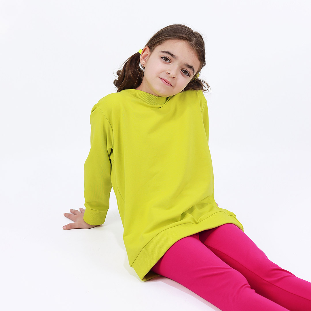 Joyful Sweatshirt is a loose fit top with minimalist design in bright lime colour that features a ribbed neck, sleeve ends and bottom hem. Front view, a girl sitting. Children, 3 -10 yrs. BonnyJoy